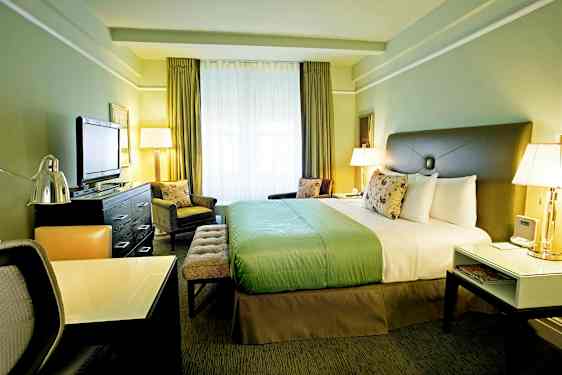 Hotellihuone, jossa on suuri sänky, työpöytä ja televisio. Huone on sisustettu vihrein sävyin ja siinä on suuret ikkunat. (Tekoälyn luoma teksti)