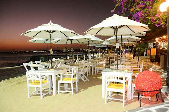 Valkoiseksi maalattuja pöytiä ja tuoleja suurilla varjoilla rannalla hämärän aikaan. (Tekoälyn luoma teksti)