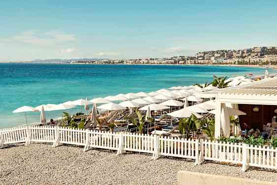 Aurinkoinen ranta, sininen meri, aurinkovarjoja ja istumapaikkoja valkoisen aidan ympäröimänä. (Tekoälyn luoma teksti)