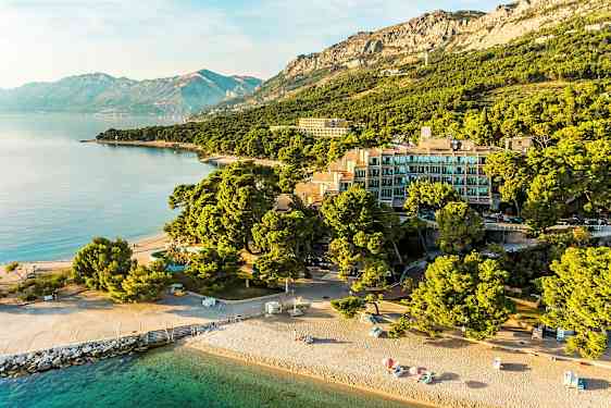 Ilmakuva rannasta, jossa on aurinkovarjoja, rakennuksia puiden keskellä ja vuoria taustalla. (Tekoälyn luoma teksti)