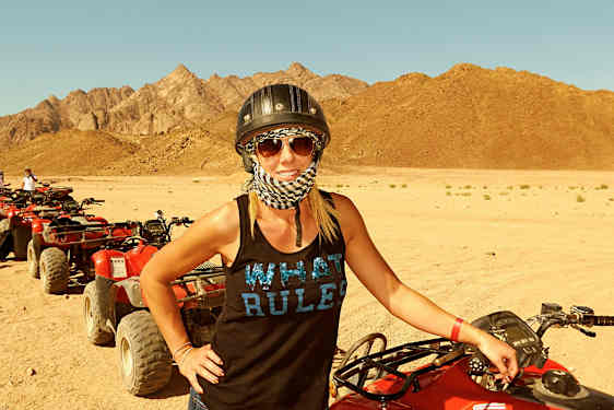 Nainen seisoo punaisen mönkijän vieressä hiekkasella autiomaalla, taustalla näkyy vuoria. (Tekoälyn luoma teksti)