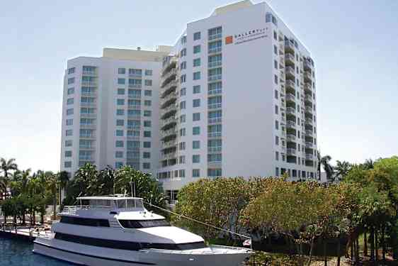 Kuva suuresta valkoisesta rakennuksesta, jossa on useita kerroksia ja parvekkeita. Rakennuksen edessä näkyy valkoinen moottorivene kiinnitettynä veden äärellä sekä puita ja vihreää kasvillisuutta. (Tekoälyn luoma teksti)