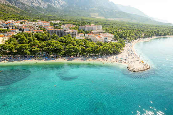 Ilmakuva rannasta, jossa kirkas sininen vesi, aurinkotuoleja ja tiivis asutus metsän ja vuorten ympäröimänä. (Tekoälyn luoma teksti)