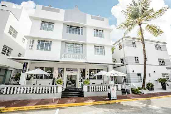 Valkoinen modernisti suunniteltu hotelli, jossa on suuret ikkunat ja kuisti aurinkovarjoilla. (Tekoälyn luoma teksti)