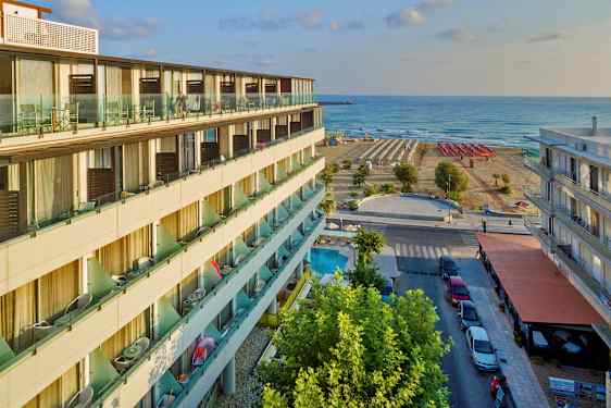 Hotellirakennus parvekkeineen, katu pysäköintipaikkoineen ja jalkakäytävineen sekä ranta aurinkotuoleineen ja meri taustalla. (Tekoälyn luoma teksti)