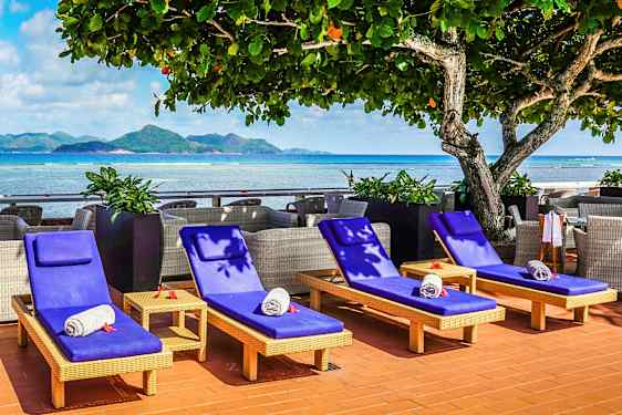 Aurinkotuolit sinisillä tyynyillä ja rullatuilla pyyhkeillä sijaitsevat meren rannalla, taustalla vuoria. (Tekoälyn luoma teksti)