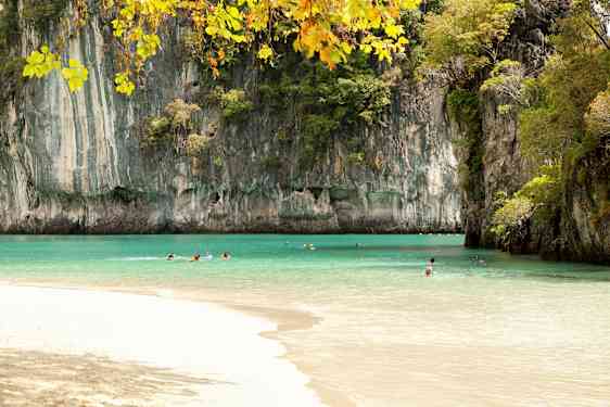 Kuvassa on ranta, jonka kirkkaan turkoosi vesi on korkeiden kallioiden ympäröimä ja kasvien peittämä. Useita henkilöitä ui vedessä kirkkaan taivaan alla. (Tekoälyn luoma teksti)