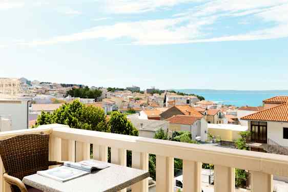 Parveke, jossa pöytä ja tuoli, näkymä kaupungille, jossa punaisia kattoja, ja taustalla meri. (Tekoälyn luoma teksti)