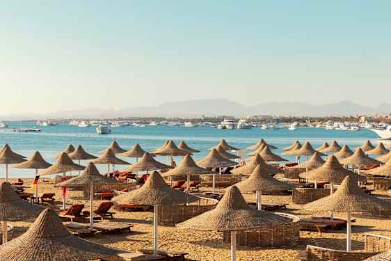 Ranta, jossa on aurinkotuoleja ja luonnonmateriaaleista valmistettuja aurinkovarjoja, taustalla meri ja veneitä. (Tekoälyn luoma teksti)