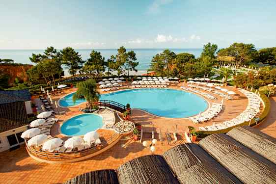 Näkymä uima-altaalle, jonka ympärillä on aurinkotuoleja ja aurinkovarjoja, viheralueiden ympäröimänä ja meri taustalla. (Tekoälyn luoma teksti)