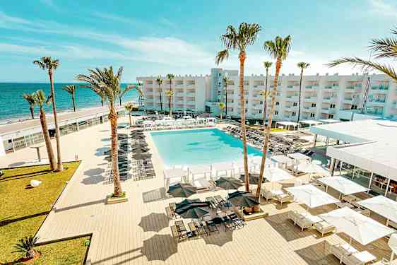 Näkymä lomakeskuksesta, jossa on suuri uima-allas, ympäröity aurinkotuoleilla ja -varjoilla, palmuja sekä rakennus taustalla lähellä merta. (Tekoälyn luoma teksti)