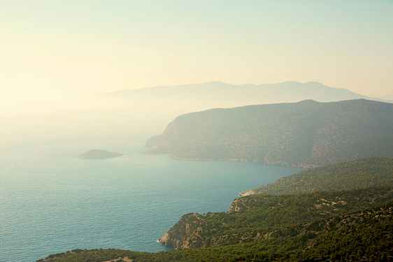 Rannikko, jossa on jyrkkiä kallioita ja vihreitä metsiä, jotka laskeutuvat mereen. Maisemaa peittää utu. (Tekoälyn luoma teksti)