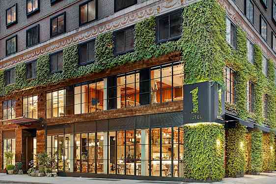 Hotellin julkisivu, jossa on muratin peittämä seinä ja suuret lasi-ikkunat, joista näkyy sisätilojen lämmin valaistus. (Tekoälyn luoma teksti)