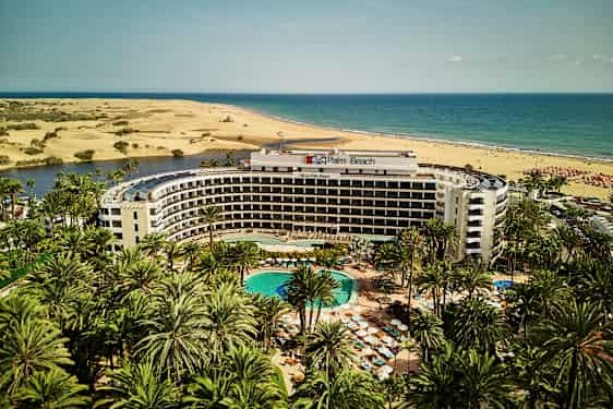 Palm Beach hotelli, jonka edessä on uima-allas, ympäröitynä palmuilla, taustalla hiekkadyynit ja meri. (Tekoälyn luoma teksti)