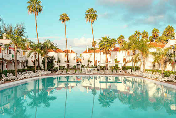 Hotellialue, jossa on valkoiseksi rapattuja rakennuksia punaisilla katoilla, ympäröity palmuilla, ja suuri uima-allas aurinkotuoleilla. (Tekoälyn luoma teksti)