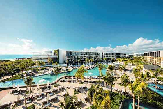 Nykyaikainen hotellialue, jossa on useita uima-altaita, vehreää kasvillisuutta ja palmuja, sijaitsee rannikolla ja tarjoaa näkymän merelle kirkkaan sinisen taivaan alla. (Tekoälyn luoma teksti)