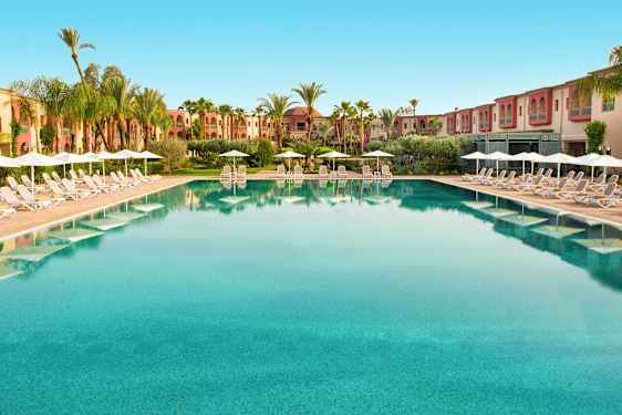 Suuri uima-allas, jonka reunoilla on aurinkotuoleja ja sateenvarjoja, ympäröity palmuilla ja rakennuksilla. (Tekoälyn luoma teksti)