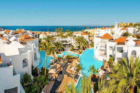 Uima-allasalue, jossa valkoiset rakennukset ja punaiset katot, ympäröity palmuilla ja näkymä merelle. (Tekoälyn luoma teksti)