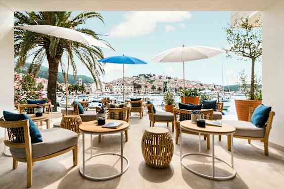 Ulkoterassi, jossa pöytiä ja tuoleja varjojen alla, ympärillä palmuja ja näkymä viehättävään satamaan ja kaupunkiin. (Tekoälyn luoma teksti)