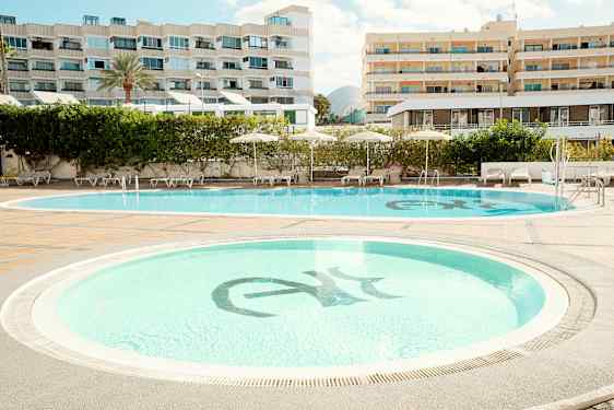 Kaksi uima-allasta, joita ympäröivät aurinkotuolit ja aurinkovarjot, taustalla hotellirakennuksia. (Tekoälyn luoma teksti)