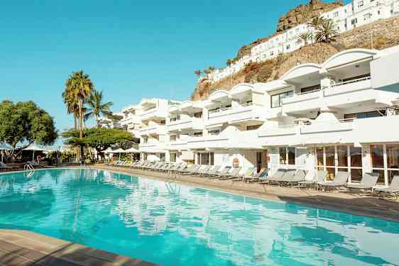 Uima-allas valkoisen hotellirakennuksen edessä, jossa on useita parvekkeita. Taustalla näkyy palmuja ja rinne. (Tekoälyn luoma teksti)