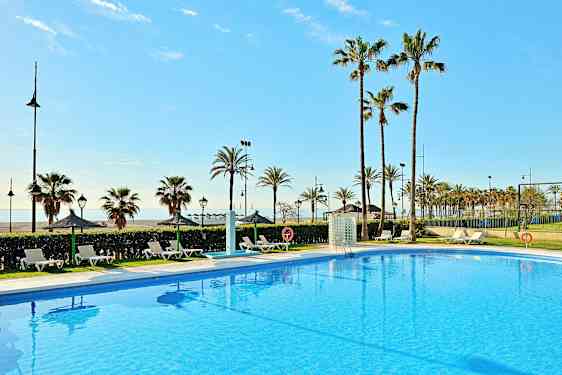 Kirkas sininen uima-allas, jonka ympärillä on aurinkotuoleja, taustalla palmuja ja kauempana meri. (Tekoälyn luoma teksti)