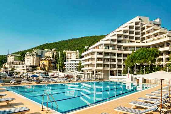 Uima-allasalue aurinkotuoleineen hotellin edessä, ympärillä vihreitä kukkuloita ja muita rakennuksia taustalla. (Tekoälyn luoma teksti)