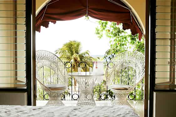 Näkymä huoneesta terassille, jossa on kaksi valkoista tuolia ja pöytä laseineen, ympäröitynä vehreydellä ja palmupuilla. (Tekoälyn luoma teksti)