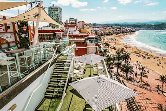 Kattoterassi, jossa pöytiä ja aurinkovarjoja, lasikaiteiden ympäröimä, näkymä rannalle ja kaupungille meren äärellä. (Tekoälyn luoma teksti)