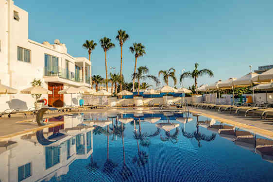 Kuva ulkouima-altaasta, jonka ympärillä on aurinkotuoleja ja aurinkovarjoja, taustalla palmuja ja valkoinen rakennus. (Tekoälyn luoma teksti)