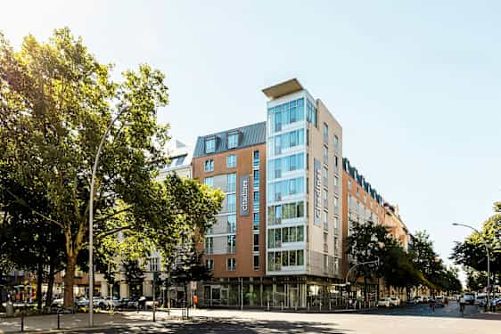 Moderni hotelli lasiseinällä, ympärillä puita ja kaupunkimaisemaa. (Tekoälyn luoma teksti)