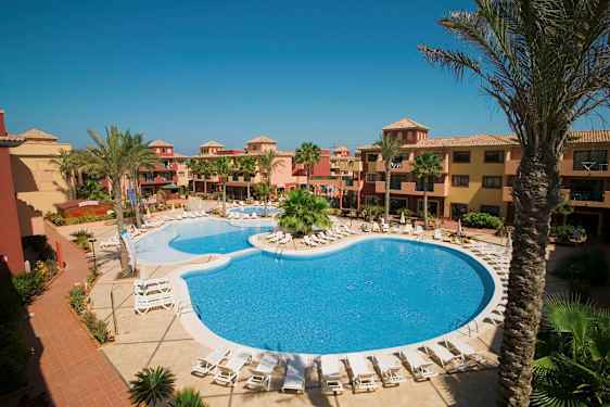 Hotellialue, jossa uima-allas ja aurinkotuoleja, ympäröity palmupuiden ja lämpimänsävyisten rakennusten kanssa. (Tekoälyn luoma teksti)