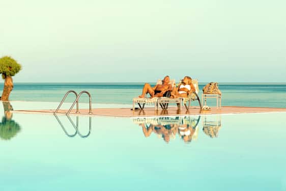 Kaksi henkilöä makaa aurinkotuoleilla uima-altaan äärellä, näkymä merelle. (Tekoälyn luoma teksti)