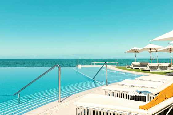 Ikkunaton uima-allas, josta on näkymä merelle, ympärillä aurinkotuoleja ja aurinkovarjoja. (Tekoälyn luoma teksti)