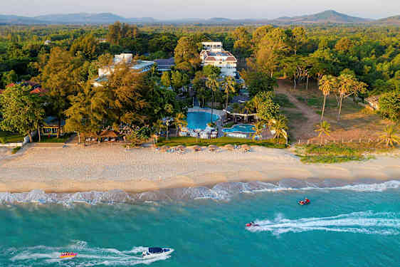 Ranta, jossa on aurinkovarjoja ja tuoleja, sekä hotellirakennus ja uima-altaita taustalla, ympäröitynä kasvillisuudella. (Tekoälyn luoma teksti)