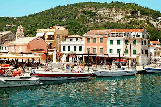 Satama, jossa useita veneitä värikkäiden rakennusten edessä, ympäröimänä mäkiä. (Tekoälyn luoma teksti)