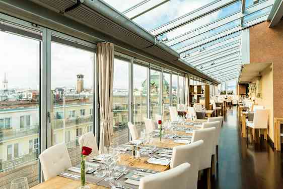 Valoisa ravintola, jossa on lasiseinät ja -katto, pöydät on katettu valkoisilla tuoleilla ja jokaiseen pöytään on asetettu ruusu. (Tekoälyn luoma teksti)