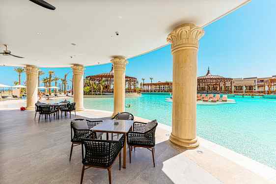 Ulkoterassialue, jossa pöytiä ja tuoleja uima-altaan lähellä, ympärillä pylväitä ja aurinkotuoleja. (Tekoälyn luoma teksti)