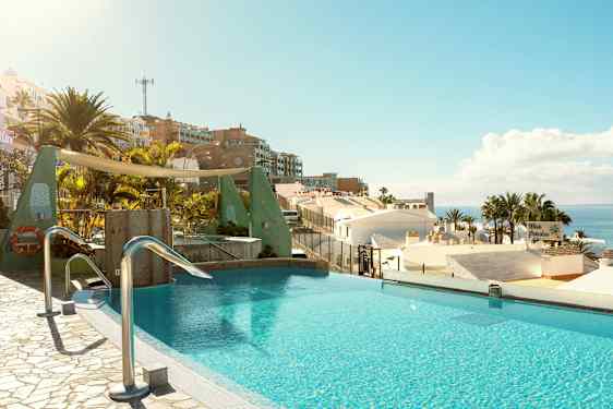 Päättymättömän näköinen uima-allas, josta on näkymä kattojen yli palmupuiden ja meren suuntaan aurinkoisessa maisemassa. (Tekoälyn luoma teksti)