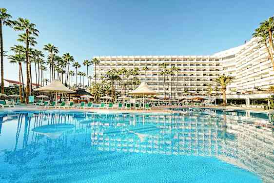 Hotellirakennus, jossa suuri uima-allas ja aurinkotuoleja etualalla, ympäröitynä palmuilla. (Tekoälyn luoma teksti)