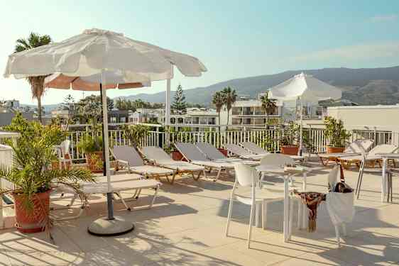 Auringonlaskuterassi, jossa on aurinkotuoleja, aurinkovarjoja, pöytiä ja tuoleja, ympäröitynä vehreydellä ja näkymä rakennuksiin ja vuorille. (Tekoälyn luoma teksti)