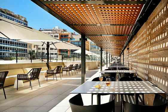 Ulkoterassi, jossa on pöytiä ja tuoleja puupergolan ja suurten aurinkovarjojen alla. (Tekoälyn luoma teksti)