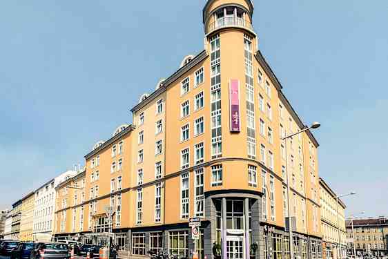 Vaaleankeltainen rakennus, jossa on useita ikkunoita, kyltti ja pyöreä kulmajulkiin. (Tekoälyn luoma teksti)