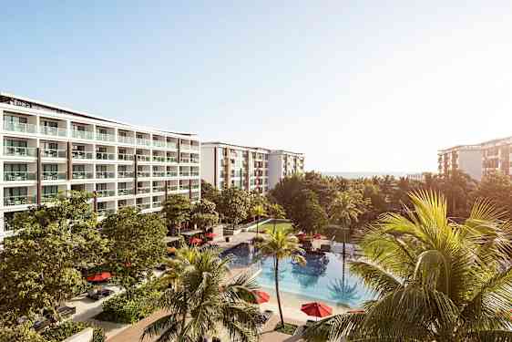 Hotellikompleksi, jossa on useita kerroksia ja parvekkeita, vehreiden puiden ympäröimänä. Rakennusten edessä on ulkouima-allas, jossa on punaisia aurinkovarjoja. (Tekoälyn luoma teksti)