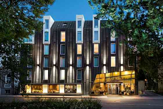 Nykyaikaisen hotellin julkisivu, jossa on lasipinta ja valaistuja ikkunoita, ympärillä puita. (Tekoälyn luoma teksti)