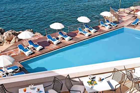 Uima-allas, jossa aurinkotuoleja ja päivänvarjoja puuterassilla meren äärellä. Etualalla näkyy pöytä tuoleineen ja juomineen. (Tekoälyn luoma teksti)