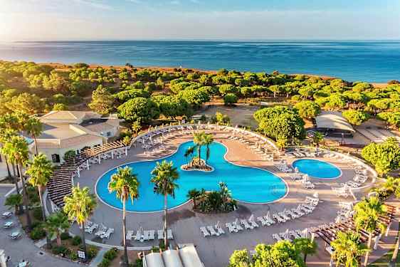 Ilmakuva hotellialueesta, jossa on suuria uima-altaita, aurinkotuoleja ja ympäröivää vehreyttä lähellä rantaa. (Tekoälyn luoma teksti)