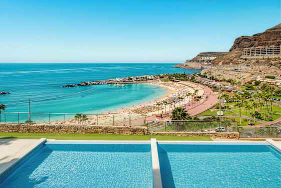 Kuva rannasta, jossa aurinkovarjoja, turkoosia vettä ja uima-allas etualalla. (Tekoälyn luoma teksti)