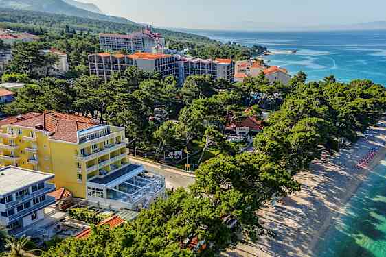 Ilmakuva rannasta, jossa on aurinkotuoleja ja aurinkovarjoja hotellirakennusten ja kasvillisuuden vieressä. (Tekoälyn luoma teksti)