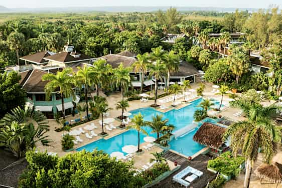 Ilmakuva hotellialueesta, jossa on suuri uima-allas, jota ympäröivät aurinkotuolit ja aurinkovarjot sekä vehreitä puita ja kasvillisuutta. (Tekoälyn luoma teksti)